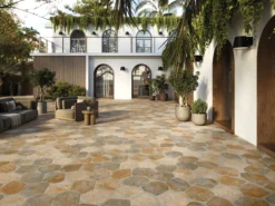 Open-air courtyard with beige stone-tiled floor, wicker seating, and potted plants against a white wall with arched windows and ivy climbing overhead.