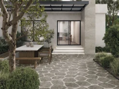 Modern courtyard with a stone-paved patio, wooden dining table and benches, and a large tree beside a concrete house with sliding glass doors.