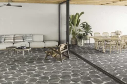 Open-concept living and dining area with a stone-patterned floor, beige sofa with patterned cushions, and a long wooden dining table with rattan chairs under wooden ceiling beams.