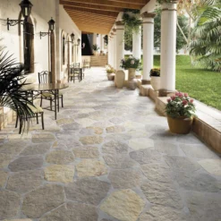 A sunlit stone-covered veranda with white columns, hanging lanterns, and wrought-iron chairs along a colonnade led to outdoor stairs, surrounded by potted plants and greenery.
