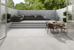 Modern outdoor patio with a grey sectional sofa on a raised platform and a round dining table with chairs to the right, surrounded by stone walls and greenery.