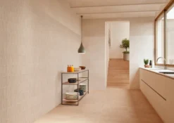 Minimalist kitchen with light beige tiles, a metal shelf holding plates and bottles, and stairs leading to an upper level with plants nearby.