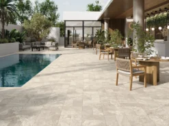 Modern outdoor patio with a rectangular pool on the left, lounge seating, and wooden dining tables under a covered area with greenery and glass doors in the background.