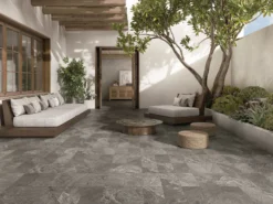 Modern outdoor patio with beige cushioned seating, wooden frames, and a large tree beside a doorway.