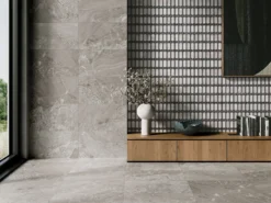 Modern interior showing large gray stone floor tiles and a black-gray geometric tiled wall with a wooden cabinet and decorative vase of white blossoms on the countertop edge.