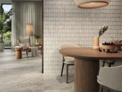 Open-concept dining area with a round wooden table, beige chairs, and a geometric textured wall; plants and daylight visible outside the glass door.