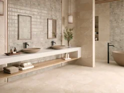 Double sink bathroom with beige stone countertops, two vessel sinks, mirrors, and a wooden bench with folded towels.